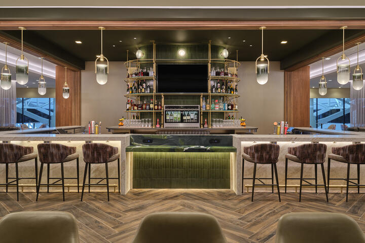Front view of lounge bar with high‑back stools, illuminated shelving, pendant lights and wood‑pattern flooring