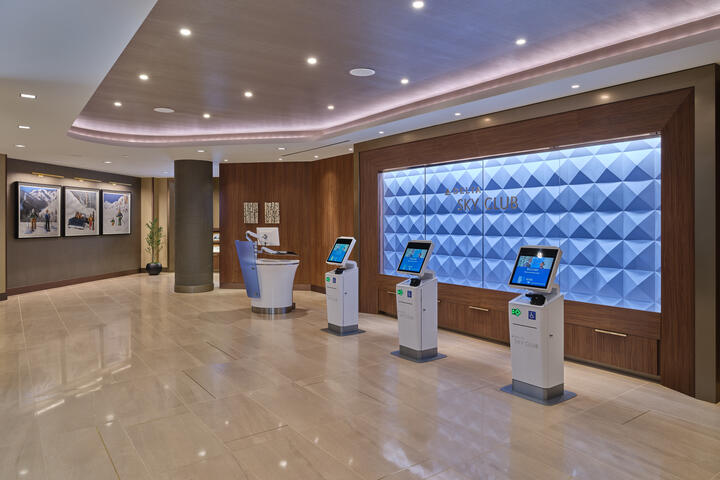 Denver Sky Club entrance with self‑service kiosks, a textured blue wall behind the Sky Club sign and a reception desk in a spacious, well‑lit lobby
