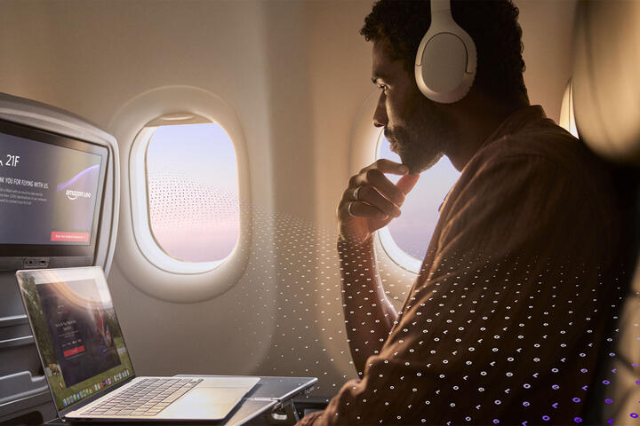 A passenger seated by an airplane window uses a laptop and wears over‑ear headphones, with an in‑flight entertainment screen visible and a subtle digital pattern overlay.