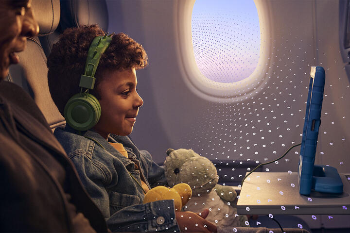 A child seated on an airplane uses a tablet while wearing headphones and holding a stuffed toy, with another seated adult and an airplane window visible, overlaid with a subtle digital pattern.