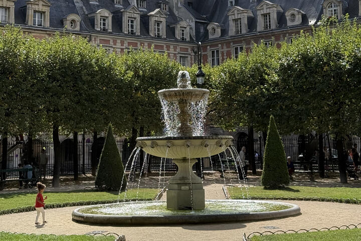 Stone fountain in a leafy Paris square, framed by manicured trees and historic red-brick buildings with slate roofs.