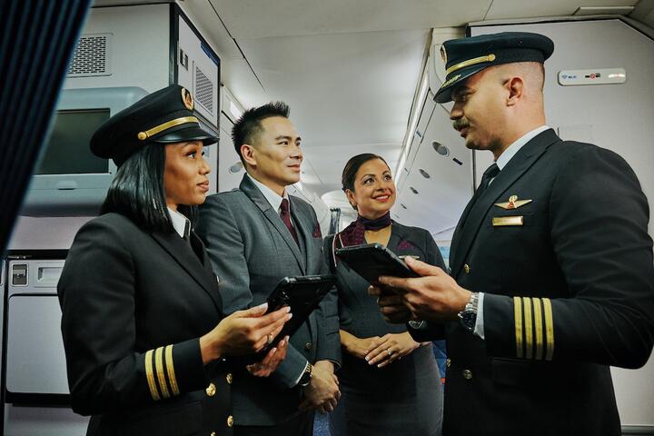 Two Delta pilots and two Delta flight attendants stand together in an airplane aisle, holding tablets and talking.