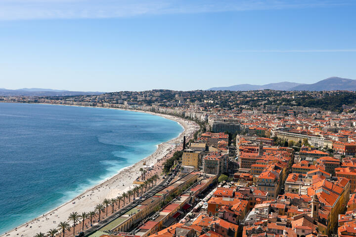 Aerial view of a curved Mediterranean beach and waterfront city with red-tiled roofs.