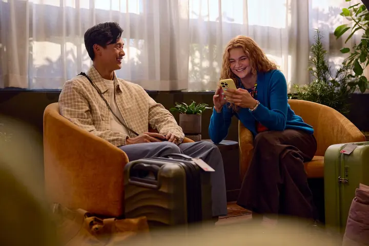 Two travelers sit in lounge chairs with rolling suitcases nearby, one looking at a smartphone, in a softly lit indoor waiting area with plants and sheer curtains.