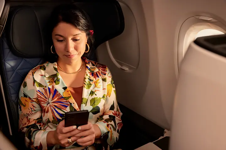Passenger seated by an airplane window holds a smartphone while sitting in a premium cabin seat, with soft lighting and aircraft interior visible.