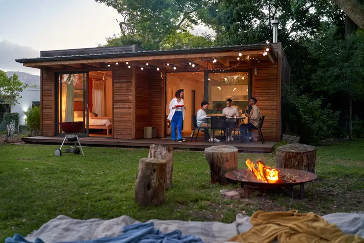 People dining outside a modern wooden cabin at dusk, with string lights, a fire pit and trees surrounding the yard.