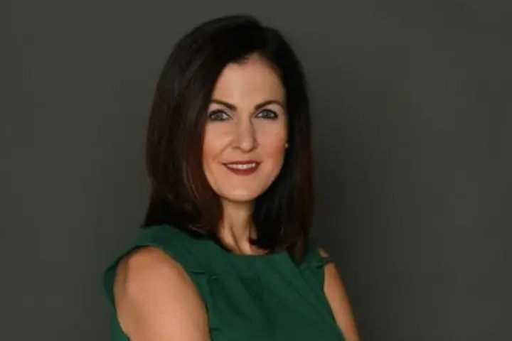 Studio portrait of a person with shoulder-length dark hair, wearing a sleeveless green dress and standing with arms crossed against a plain gray background.