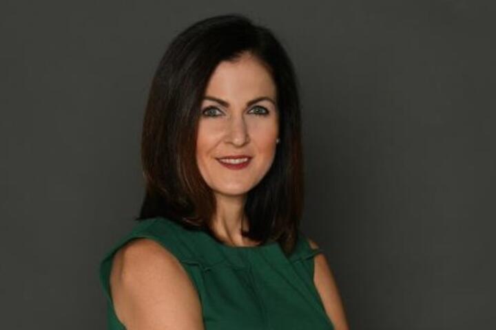 Studio portrait of a person with shoulder-length dark hair, wearing a sleeveless green dress and standing with arms crossed against a plain gray background.
