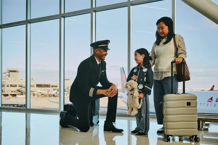 Delta pilot kneeling beside a young child holding a stuffed animal at an airport gate, while an adult stands nearby with rolling luggage; airplane visible through large windows in the background.