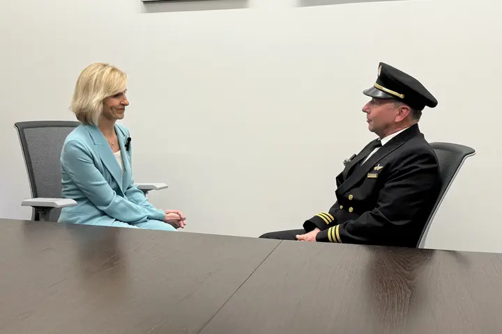 Allison Ausband and Tom Cohan sit facing each other at a conference table, Allison in a business suit and Tom in a pilot uniform.