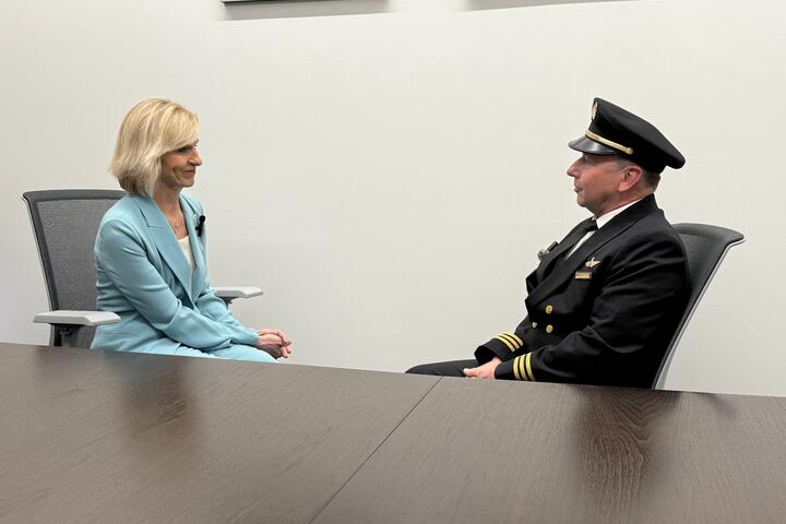 Allison Ausband and Tom Cohan sit facing each other at a conference table, Allison in a business suit and Tom in a pilot uniform.