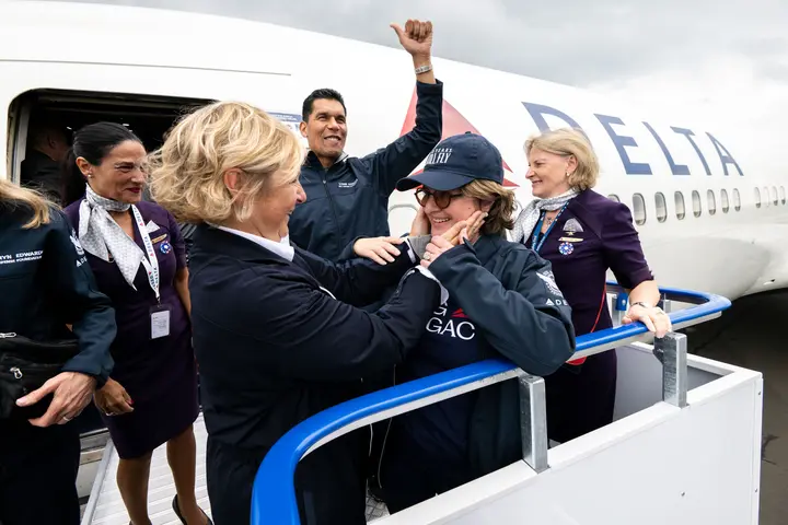 Delta employees and volunteers stand on an airport boarding ramp beside a Delta aircraft, welcoming a passenger as they exit the plane.