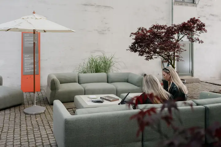 Outdoor courtyard seating area with modular sofas and a low table under an umbrella, where two people sit and talk beside plants and an event information sign.