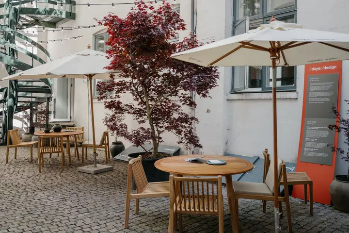 Outdoor courtyard with wooden tables and chairs under umbrellas, string lights overhead, and a red-leaf tree beside a sign for “3daysofdesign” near a building wall.