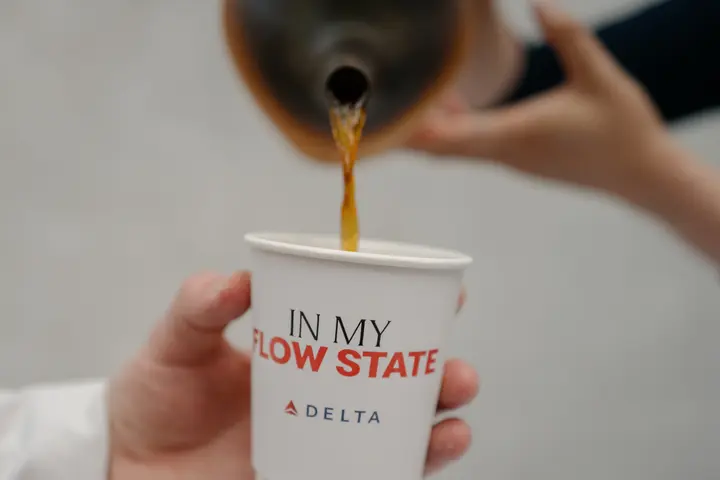Coffee being poured from a dark ceramic pitcher into a white paper cup printed with “In My Flow State” and the Delta logo.