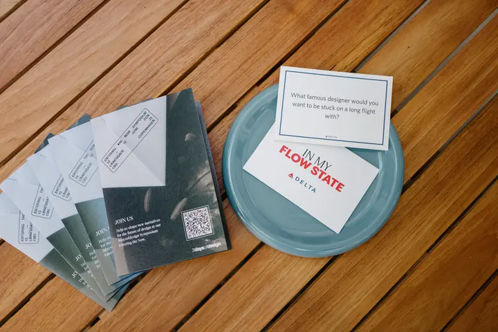 Printed prompt cards and brochures arranged on a wooden table, including a card reading “What famous designer would you want to be stuck on a long flight with?” beside an “In My Flow State” Delta card.