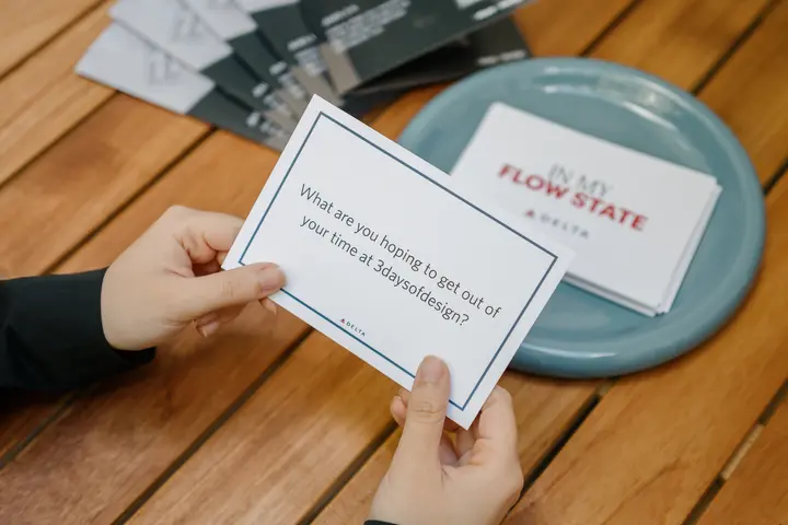 Hands holding a prompt card over a wooden table beside a bowl of cards, reading “What are you hoping to get out of your time at 3daysofdesign?”