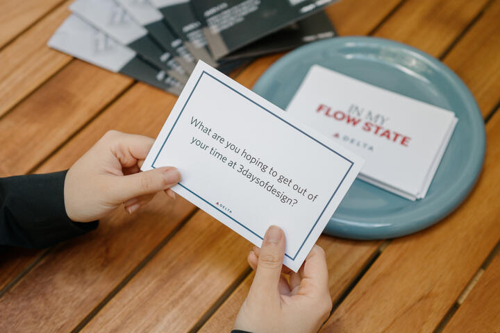 Hands holding a prompt card over a wooden table beside a bowl of cards, reading “What are you hoping to get out of your time at 3daysofdesign?”