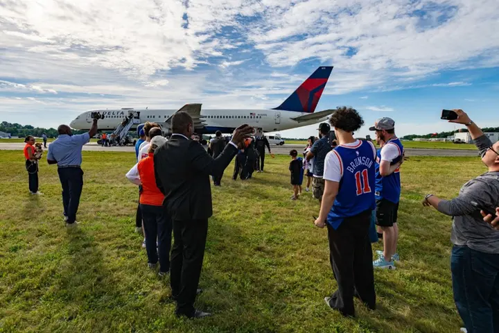 Spectators stand on grass watching a Delta airplane taxi on the runway at an airport.