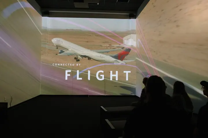 Silhouetted audience in a dark room viewing a multi-wall projection of a plane taking off, with the text “CONNECTED BY FLIGHT” overlaid.