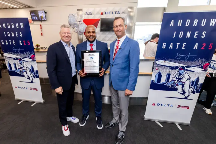 Three people pose at a Delta airport gate ceremony, with the center person holding a commemorative plaque. Banners on both sides read “Andruw Jones Gate A25,” featuring baseball-themed artwork and Delta branding.
