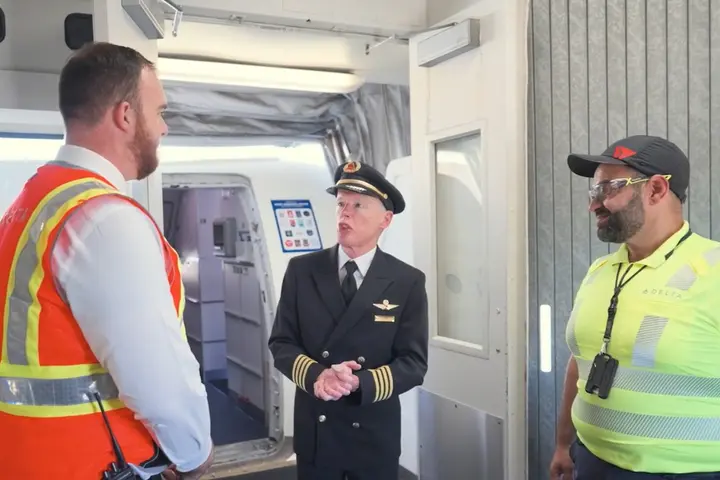 Three airport employees standing and talking near an aircraft boarding door, including a pilot in uniform and two workers wearing high-visibility safety vests.