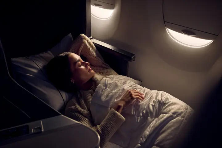  Passenger resting in a lie-flat Delta One seat with bedding, illuminated by soft cabin lighting near an aircraft window.
