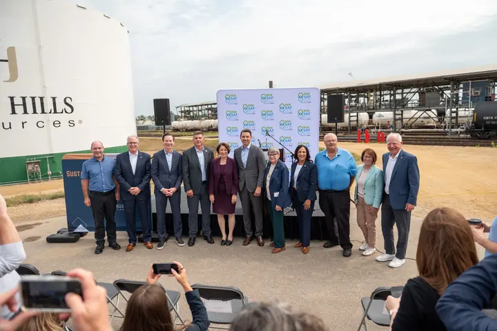 Group photo of event attendees standing in front of an industrial biofuels facility, with storage tanks, railcars, and a branded backdrop visible behind them while audience members take photos in the foreground.