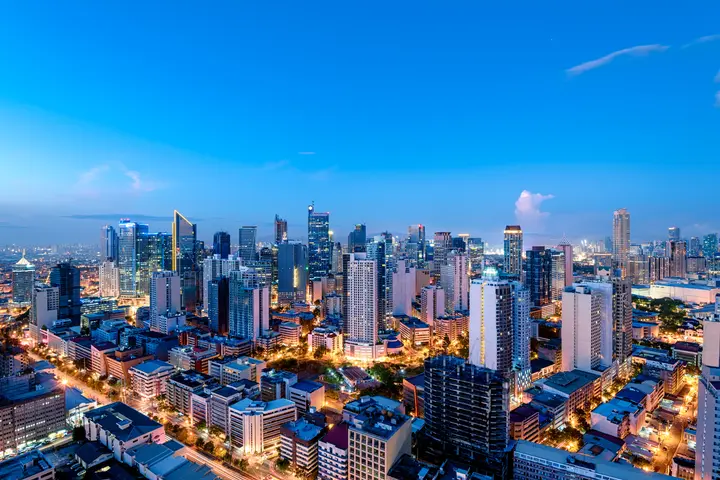 A panoramic view of a modern city skyline at dusk, with illuminated high-rise buildings, busy streets, and a bright blue evening sky.