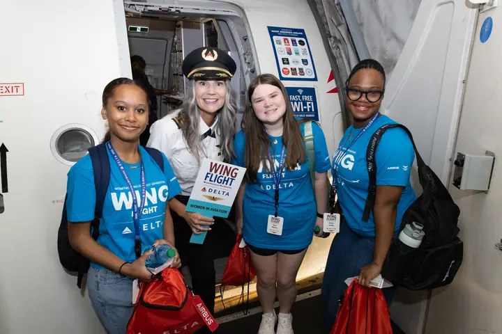 Students wearing WING Flight shirts pose with a pilot at an aircraft doorway during a Girls in Aviation Day experience.
