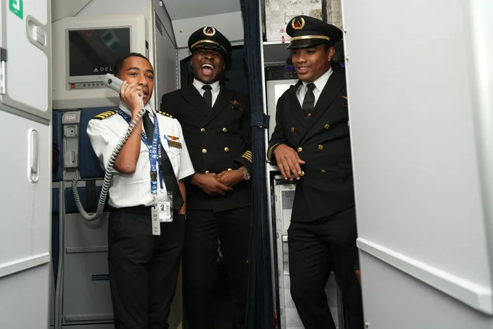 DREAM Flight participant makes an onboard announcement from the aircraft galley while Delta pilots look on.