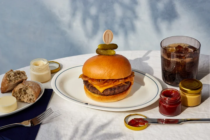Cheeseburger topped with caramelized onions and pickles, served with condiments, bread, and a soft drink on a white tablecloth.