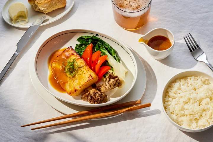 Plated glazed fish with vegetables, rice, bread, and sauce served on a white tablecloth with chopsticks and a beverage.