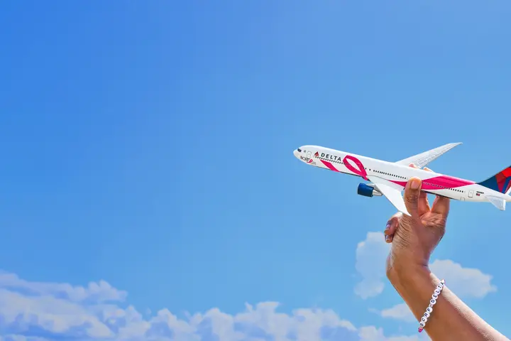 Hand holding a pink-branded Delta model airplane against a blue sky with clouds, symbolizing support for breast cancer awareness.