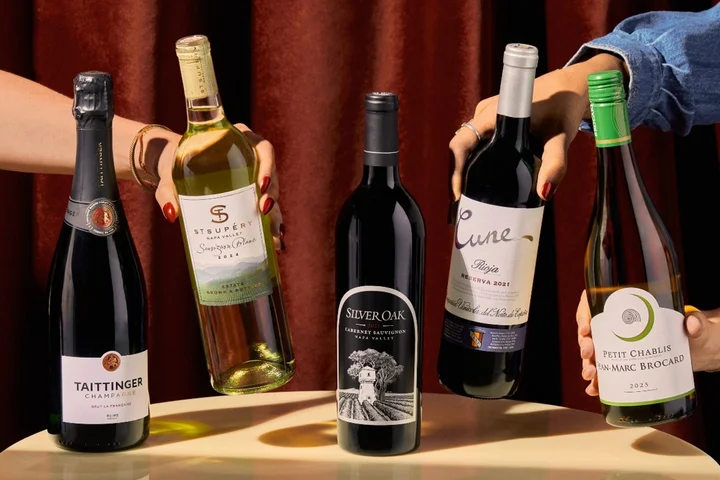 Five wine bottles on a table, with hands reaching in to hold and select different bottles against a dark red curtain backdrop.