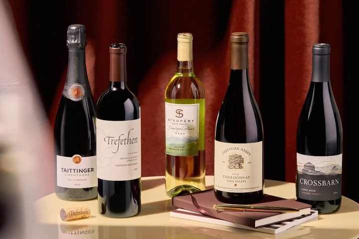 Five wine bottles arranged on a table with notebooks and a pen, set against a rich red curtain backdrop.