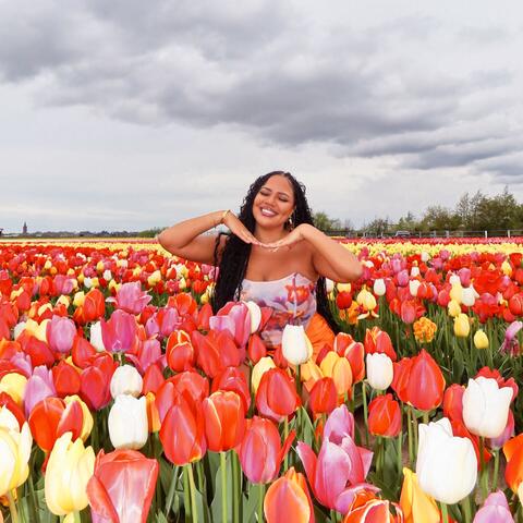 A person posing with hands raised near the shoulders while standing in a large field of colorful tulips under a cloudy sky.