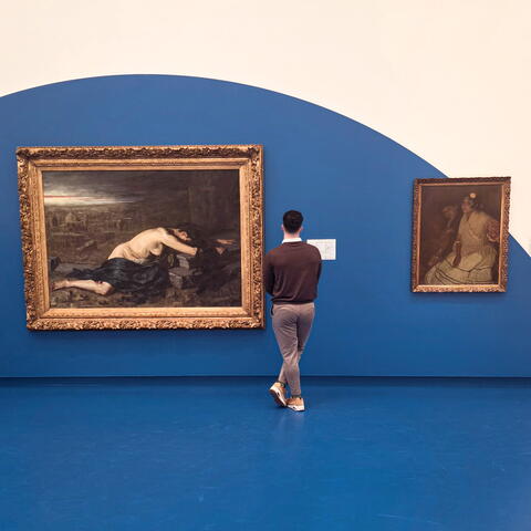 A person stands in a gallery looking at framed paintings on a blue and white wall.