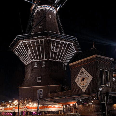 A large traditional windmill and adjacent brick brewery building are illuminated at night, with outdoor seating and lights beneath.