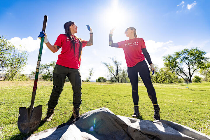 Two volunteers in red “Delta in the Community” shirts high-five while working outdoors, one holding a shovel on a sunny grassy field with trees in the background.