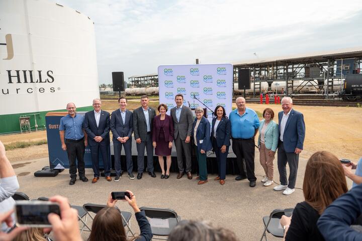 Group photo of event attendees standing in front of an industrial biofuels facility, with storage tanks, railcars, and a branded backdrop visible behind them while audience members take photos in the foreground.