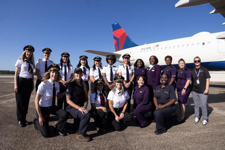Group of aviation professionals and participants posing on the tarmac in front of a Delta aircraft under a clear blue sky during a Girls in Aviation Day event.