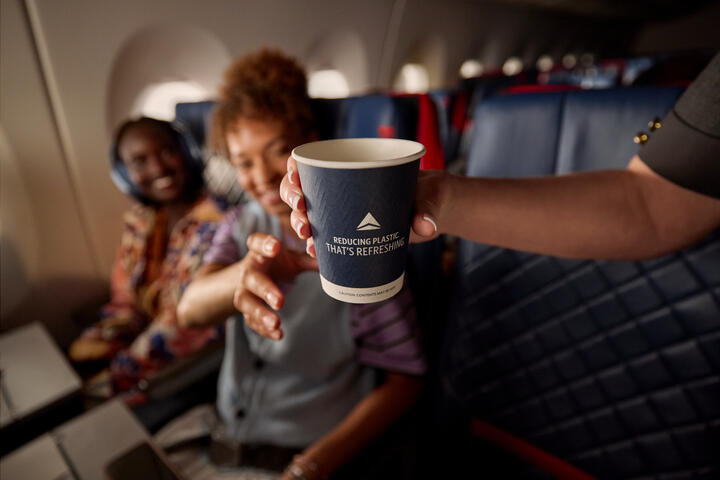 Delta's new paper cup in flight attendant's hand, ready to be handed to customer who is seated on board an aircraft and reaching for the cup.