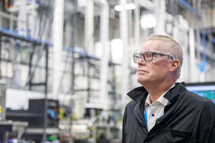 Ed Bastian wearing safety glasses standing inside an industrial facility with machinery and pipes in the background.