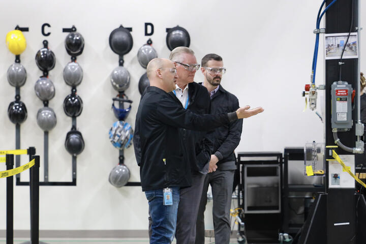 Three people wearing safety glasses review equipment inside an industrial facility.
