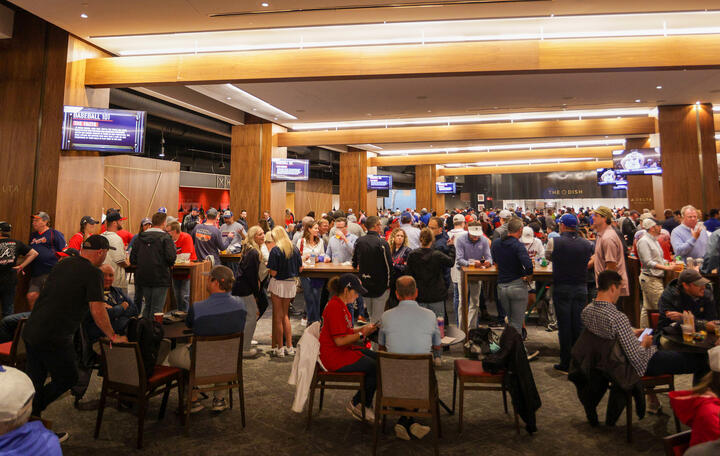 A crowded indoor stadium lounge with fans standing and sitting at tables beneath wood-paneled ceilings and multiple TV screens.