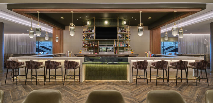 Front view of lounge bar with high‑back stools, illuminated shelving, pendant lights and wood‑pattern flooring