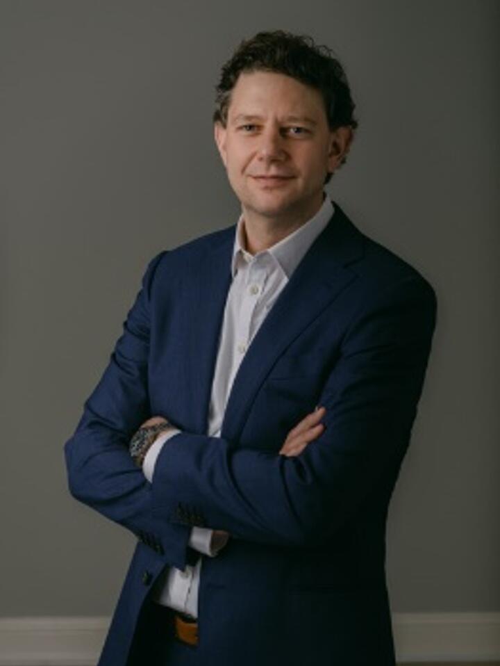 Studio portrait of a person in a dark suit and white shirt, standing with arms crossed against a plain gray background.