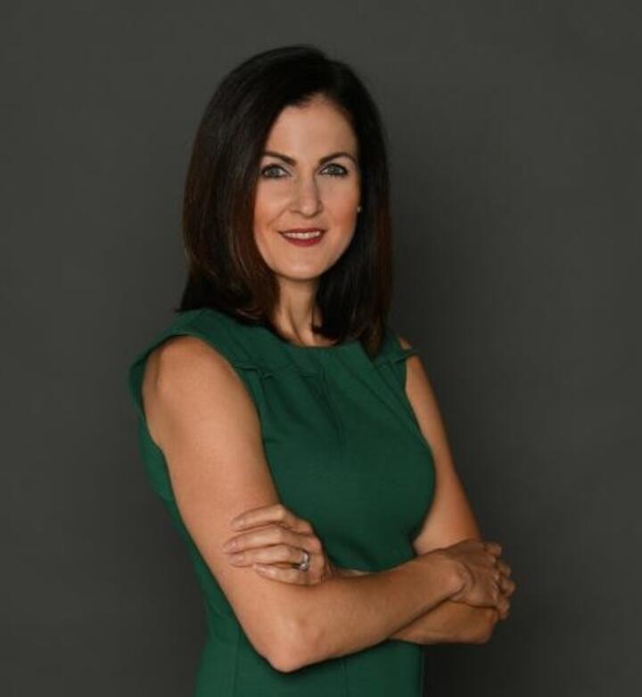 Studio portrait of a person with shoulder-length dark hair, wearing a sleeveless green dress and standing with arms crossed against a plain gray background.