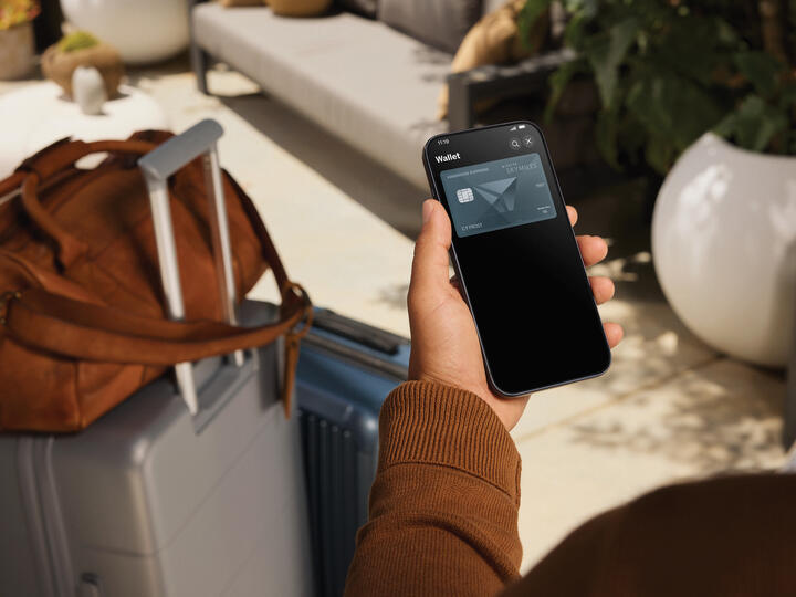 Person holding a smartphone displaying a Delta SkyMiles American Express Card in a digital wallet, with a suitcase and travel bag nearby in a lounge setting.